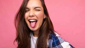 A woman wearing a blue and white flannel shirt and a white t-shirt sticking out her tongue on a pink background