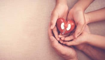 Three pairs of hands stacked on top of each other as the top pair holds a red heart-shaped block with a kidney visual painted on