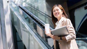 A businessperson looking up smilingly as she holds a cup of coffee in her right hand and a laptop in her left