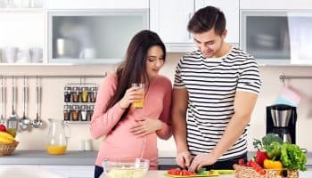 A pregnant woman looking at her husband chopping vegetables in the kitchen