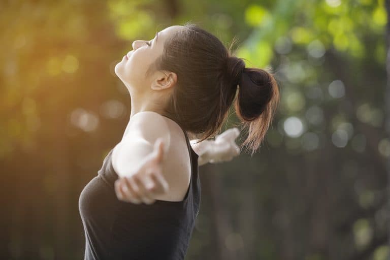 A woman in a black top raising her arms to her sides as she takes in a deep breath outdoors