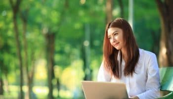 A young Asian lady is sitting on a bench and working on her laptop in a park