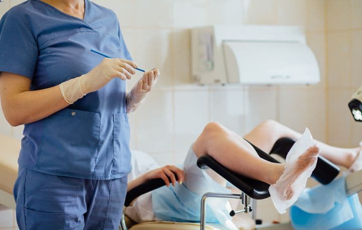 Nurse holding medical apparatuses in both hands while a woman is seen lying down with her legs on the leg rests that are connected to the chair