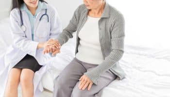 A doctor holding an elderly woman’s hand as they sit up on the edge of a bed