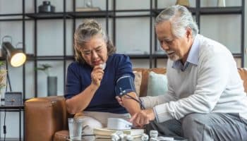 Man checking a woman’s blood pressure using a sphygmomanometer as they sit on a sofa