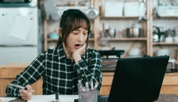 A young woman yawning while working with her laptop and notebook.