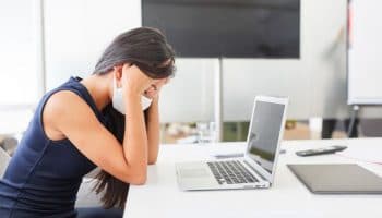 Woman wearing a white facial mask while holding her head in her hands as she sits at a table in front of her laptop.