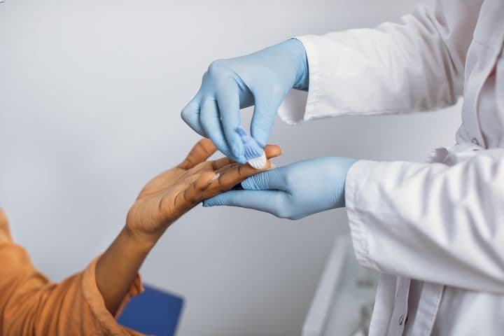 A health worker’s pair of gloved hands pricking a woman’s finger for blood testing.