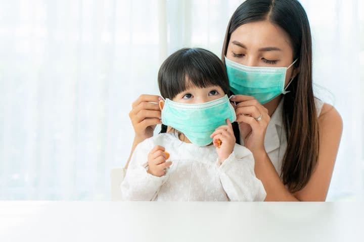 A masked mother helping her daughter to put on a mask.