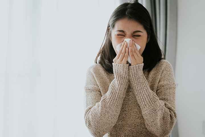 Woman in a wool sweater sneezing into a tissue that she’s holding with both hands