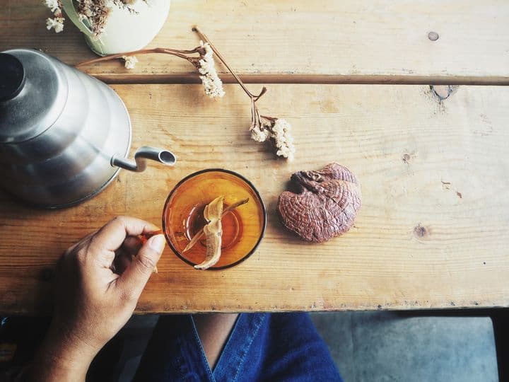 An aerial view of a woman’s hand holding a cup of tea on a wooden table, surrounded by a mushroom and teapot 