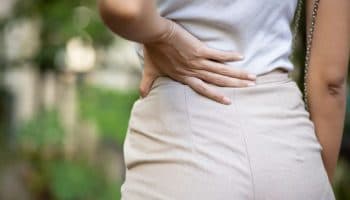 Woman holding the lower left side of her back with her left hand while standing outdoors