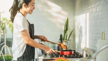 A side view of a woman in the kitchen, cooking with a pan with pieces of vegetables next to it.