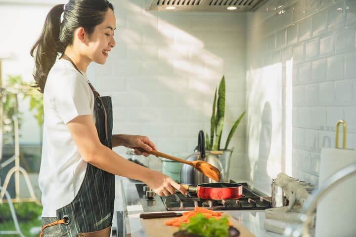 A side view of a woman in the kitchen, cooking with a pan with pieces of vegetables next to it.