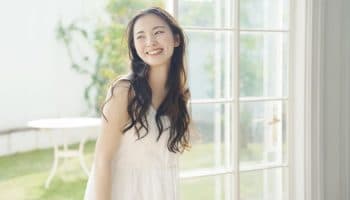 A woman smiling while standing beside a tall glass window with the view of a garden behind her.