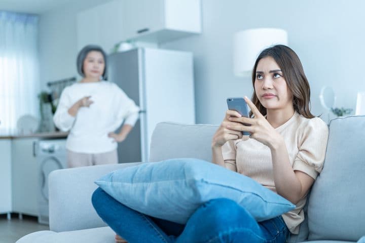 Woman holding a phone and sitting cross-legged on a sofa while rolling her eyes as she’s scolded by an elderly woman