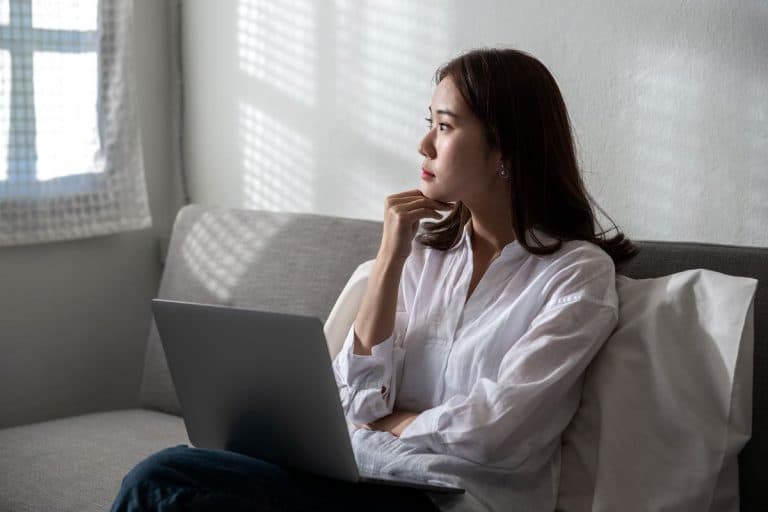 Asian woman staring out the window with her hand on her chin and a laptop on her thighs