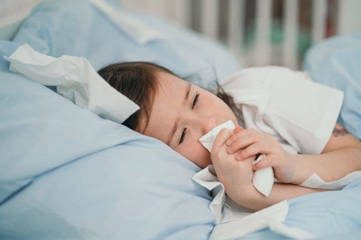 A little girl sleeping sideways on her bed, holding a tissue in front of her face