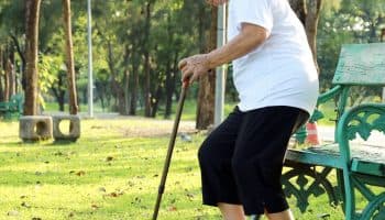 Woman grimacing in pain as she attempts to sit on a wooden park bench while holding a cane in her left hand