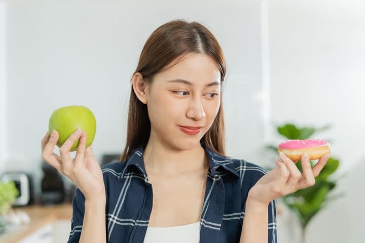 A woman is deep in thought while holding a doughnut in one hand and an apple in the other hand.