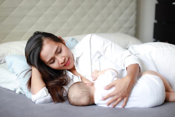 Young mother breastfeeding her newborn baby while lying on the bed.