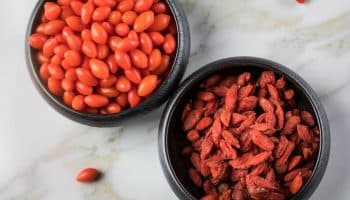 A bowl of fresh wolfberries next to a bowl of dried wolfberries on a marble surface.