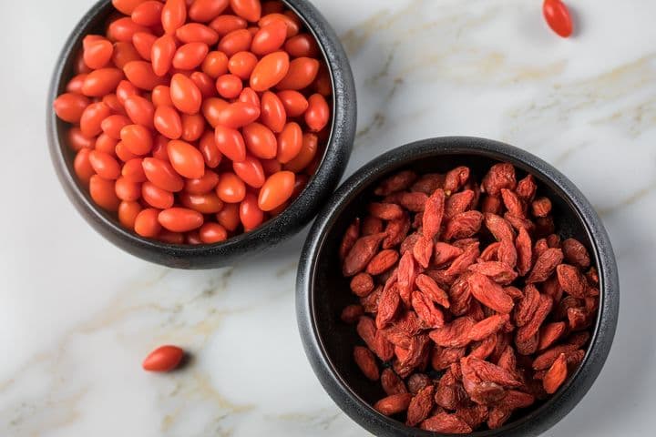 A bowl of fresh wolfberries next to a bowl of dried wolfberries on a marble surface.