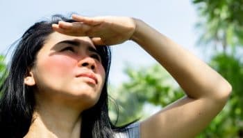 Woman standing outdoors under the sun with sunburnt cheeks covering her eyes with her left hand as she looks up