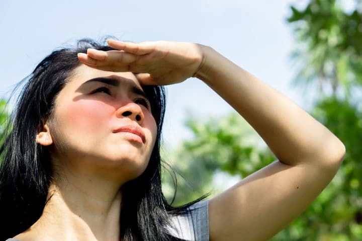 Woman standing outdoors under the sun with sunburnt cheeks covering her eyes with her left hand as she looks up