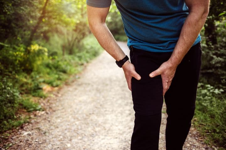 Man leaning forward as he holds his right groin area with both hands outdoors.  