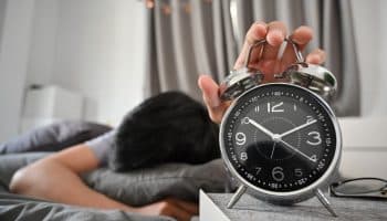 A man asleep on a bed while hugging a pillow and holding a table clock with his left hand.