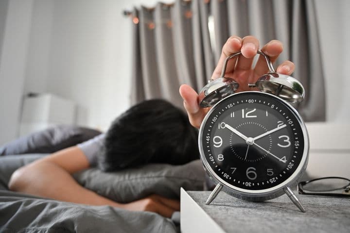 A man asleep on a bed while hugging a pillow and holding a table clock with his left hand.