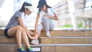 Two women sitting on stairs while one looks at the other’s ankle.