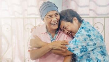 Two older women smile while hugging each other; one is a cancer patient with her head wrapped.