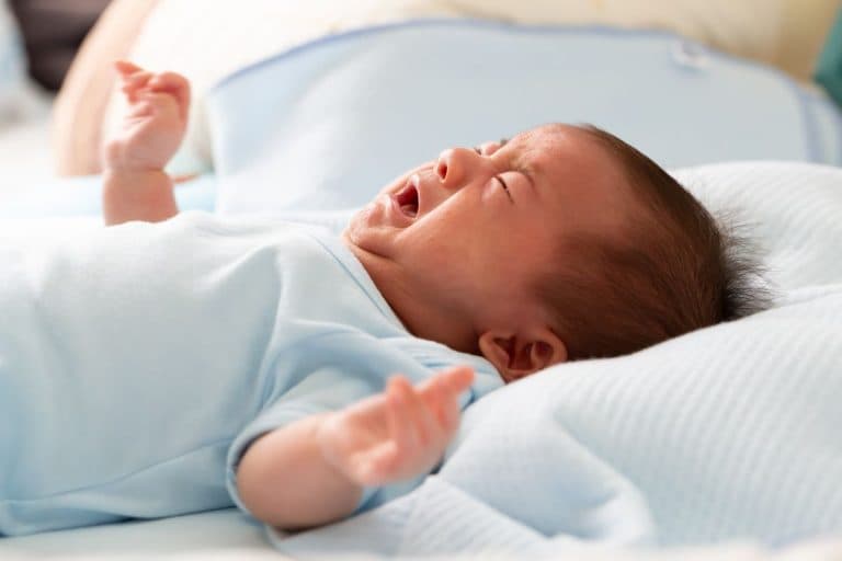 Infant crying while lying on a bed with his arms outstretched.