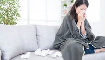 A woman wrapped in a blanket wiping her nose with a tissue while sitting on a sofa next to used tissues.