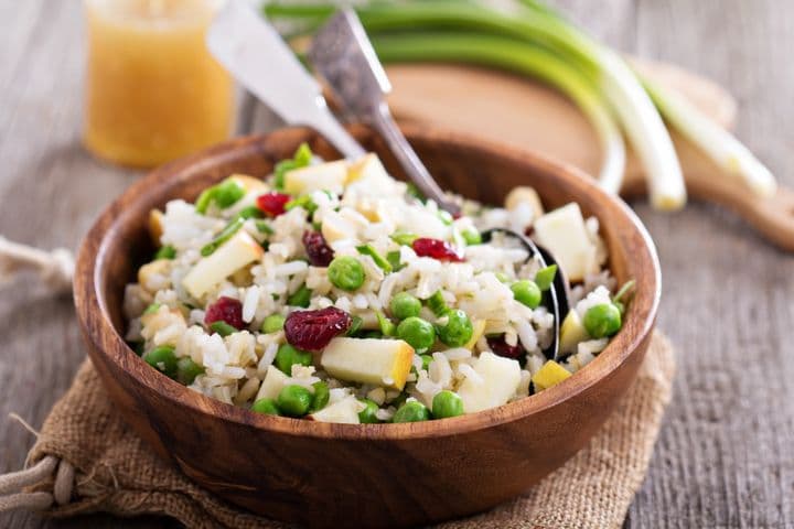 A colourful dish made of rice, tofu, peas, and cranberries served in a wooden bowl with fork and spoon.