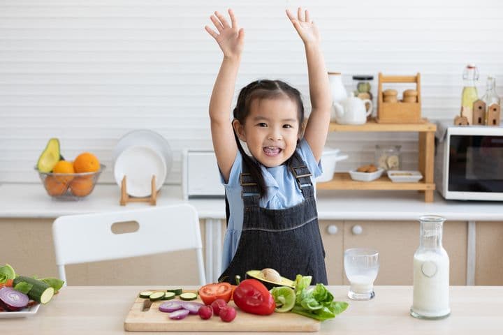 Toddler girl raises her arms and smiles standing in the kitchen in front of a cutting board filled with healthy colourful vegetables and fruits.