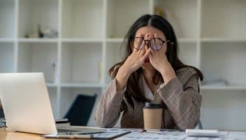 A woman covering her eyes with both hands while wearing a pair of eyeglasses as she sits a desk. 