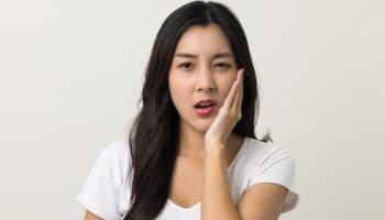 A woman grimaces in pain while holding her left cheek with her left palm.