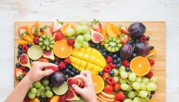 A top view of an assortment of fruits arranged on a wooden board with a child’s hands grabbing some strawberries.