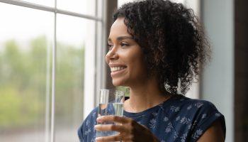 Smiling millennial biracial woman look in distance hold glass of water drink clean clear mineral aqua for body refreshment