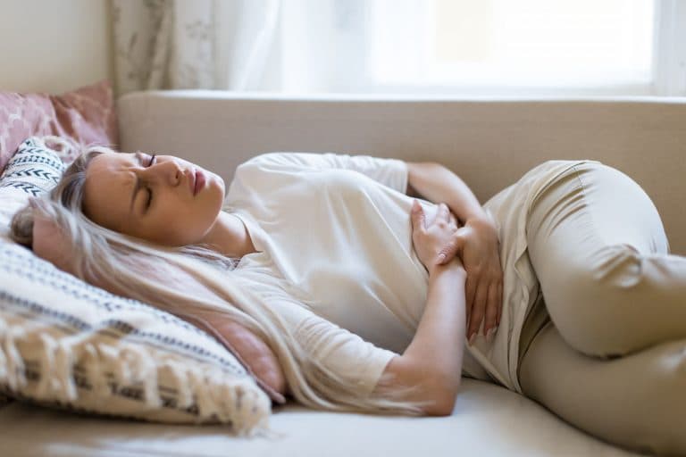 A white woman lying on the bed while holding her stomach in pain due to digestive problems