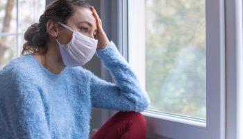 A stressed woman suffering COVID-19 wears a mask looking out the window